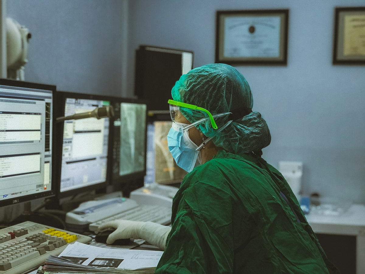 A healthcare worker in scrubs and a surgical cap at a bank of computer monitors.